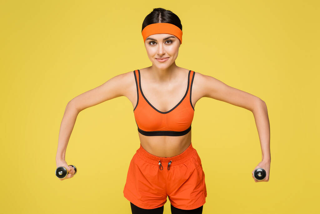 young sportswoman in orange sportswear training with dumbbells isolated on yellow - Foto, Imagen