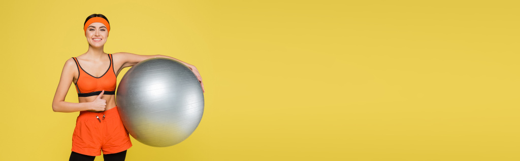 pleased sportswoman with silver-grey fitness ball showing thumb up isolated on yellow, banner - Foto, imagen