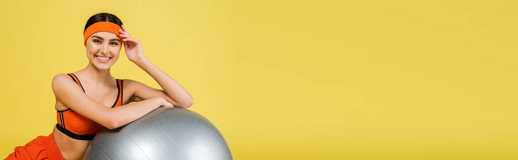 jolie sportive souriant à la caméra près du ballon de fitness isolé sur jaune, bannière - Photo, image