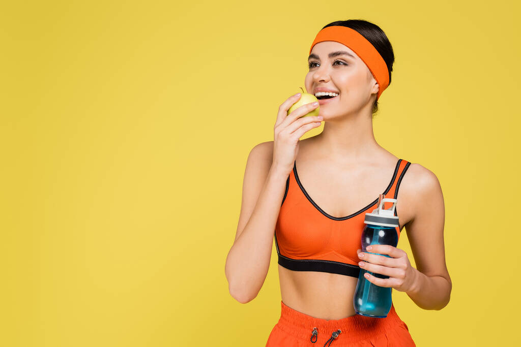 Taze elma ve spor şişesiyle mutlu sporcu kadın, sarıya izole edilmiş şekilde gülümsüyor. - Fotoğraf, Görsel