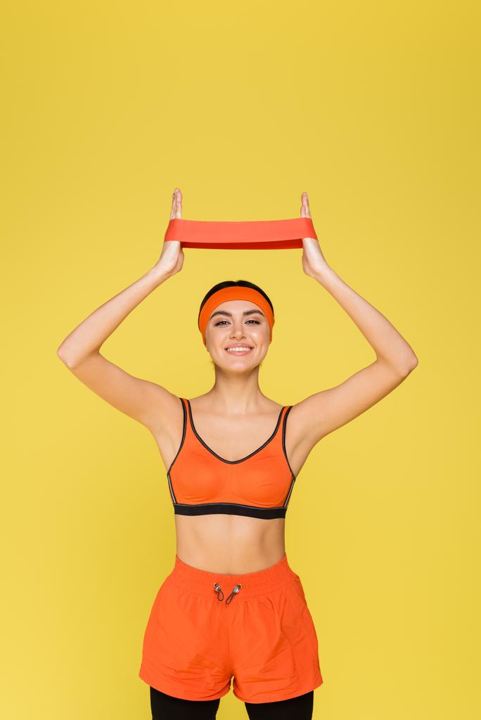 χαρούμενη γυναίκα σε πορτοκαλί αθλητικά ρούχα κατάρτισης με ζώνη αντίστασης απομονώνονται σε κίτρινο - Φωτογραφία, εικόνα