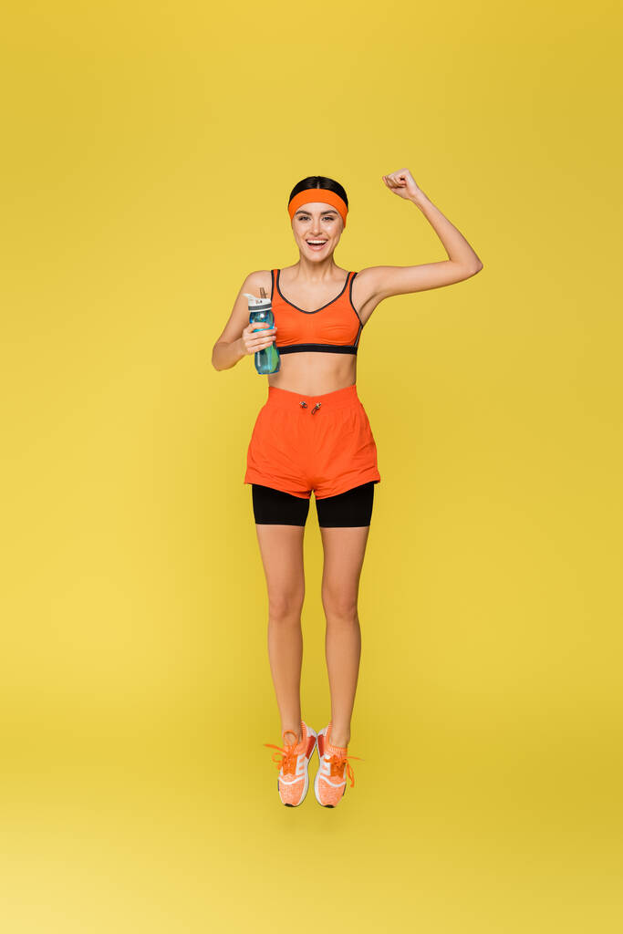 Spor şişesi atlayan neşeli sporcu kadın sarı renkte izole edilmiş zafer hareketi sergiliyor. - Fotoğraf, Görsel