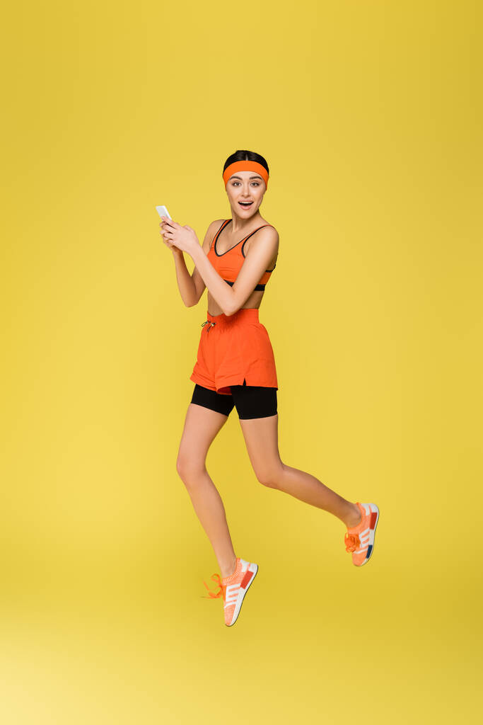 молода спортсменка стрибає під час використання смартфона ізольовано на жовтому
 - Фото, зображення
