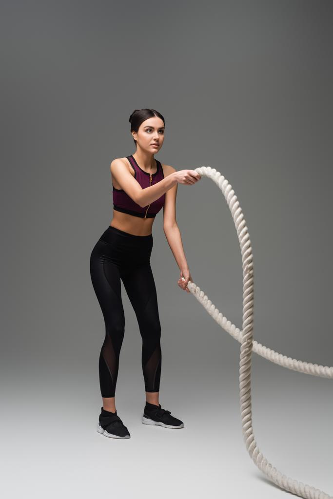 visão de comprimento total da mulher esportiva se exercitando com cordas de batalha em fundo cinza - Foto, Imagem