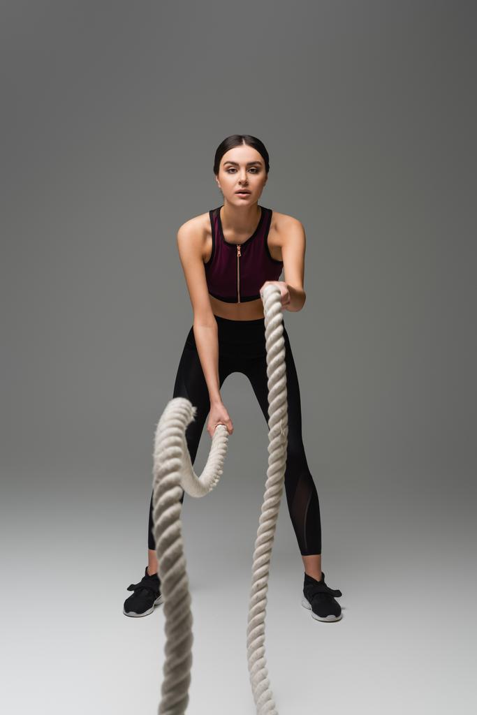 full length view of sportive woman working out with battle ropes on grey background - Foto, imagen