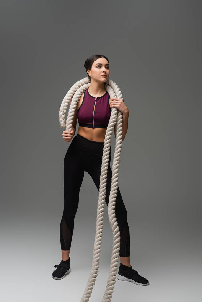 full length view of young sportswoman holding battle ropes on grey background - Photo, Image