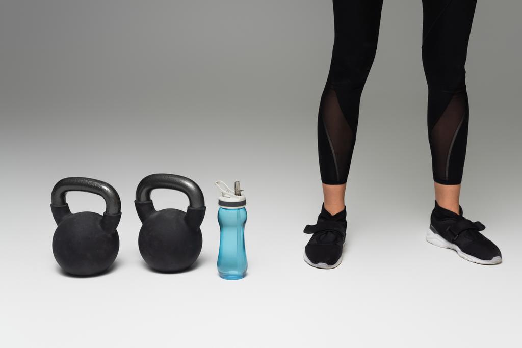 abgeschnittene Ansicht einer Sportlerin in schwarzen Leggings in der Nähe von Kettlebells und Sportflasche auf grauem Hintergrund - Foto, Bild