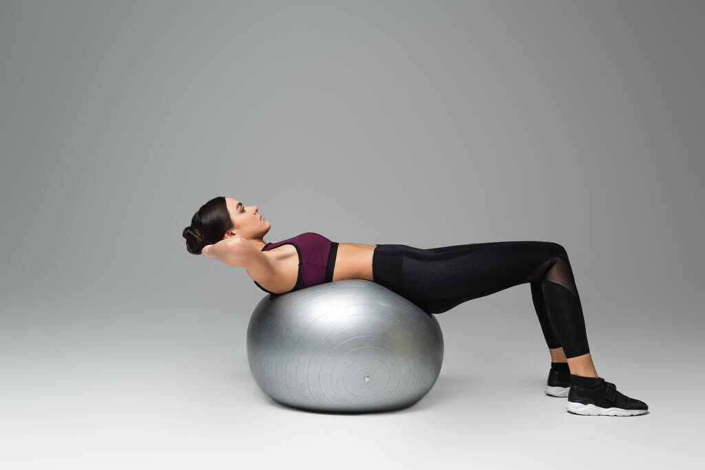 вид сбоку на женщину в черном, тренирующуюся на фитнес-мяче на сером фоне - Фото, изображение