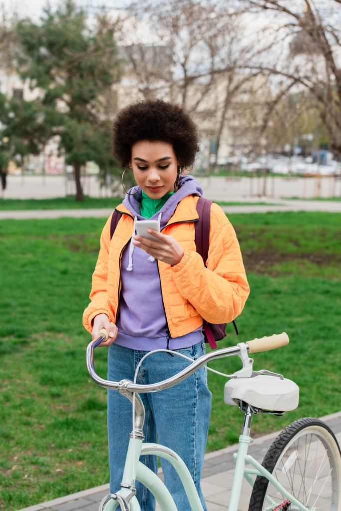 Junge Afroamerikanerin benutzt Handy, während sie in der Nähe von Fahrrad im Freien steht  - Foto, Bild