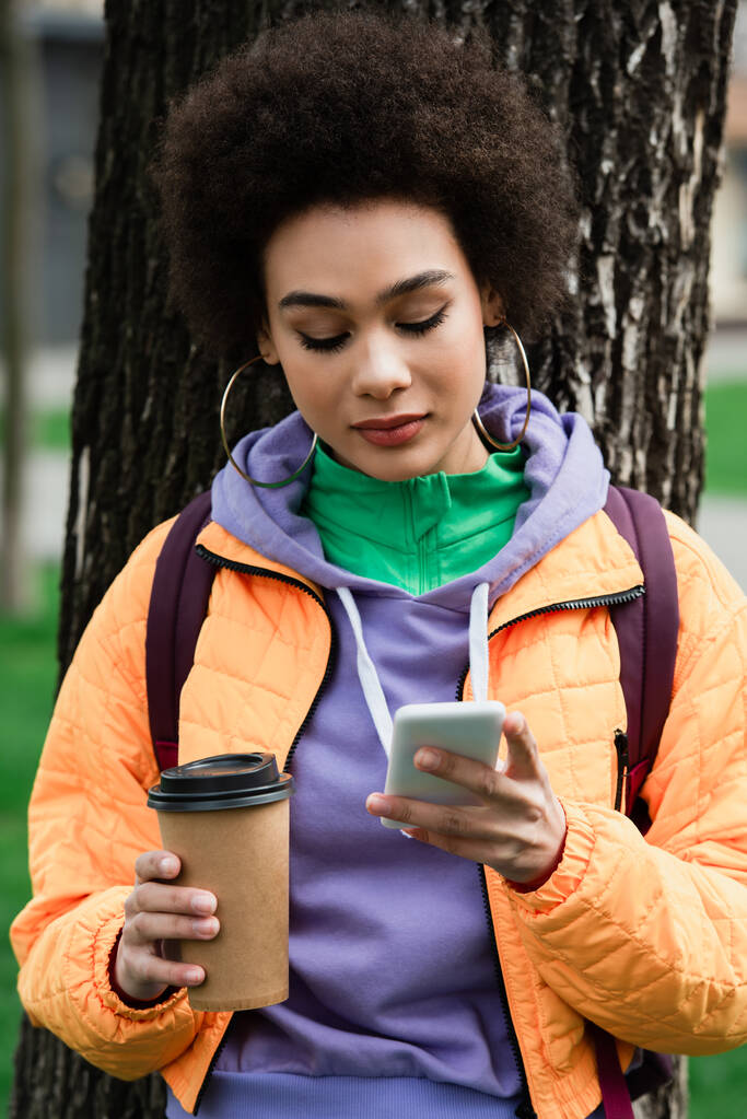 Mujer afroamericana sosteniendo una taza para llevar y usando un teléfono inteligente cerca del árbol  - Foto, imagen