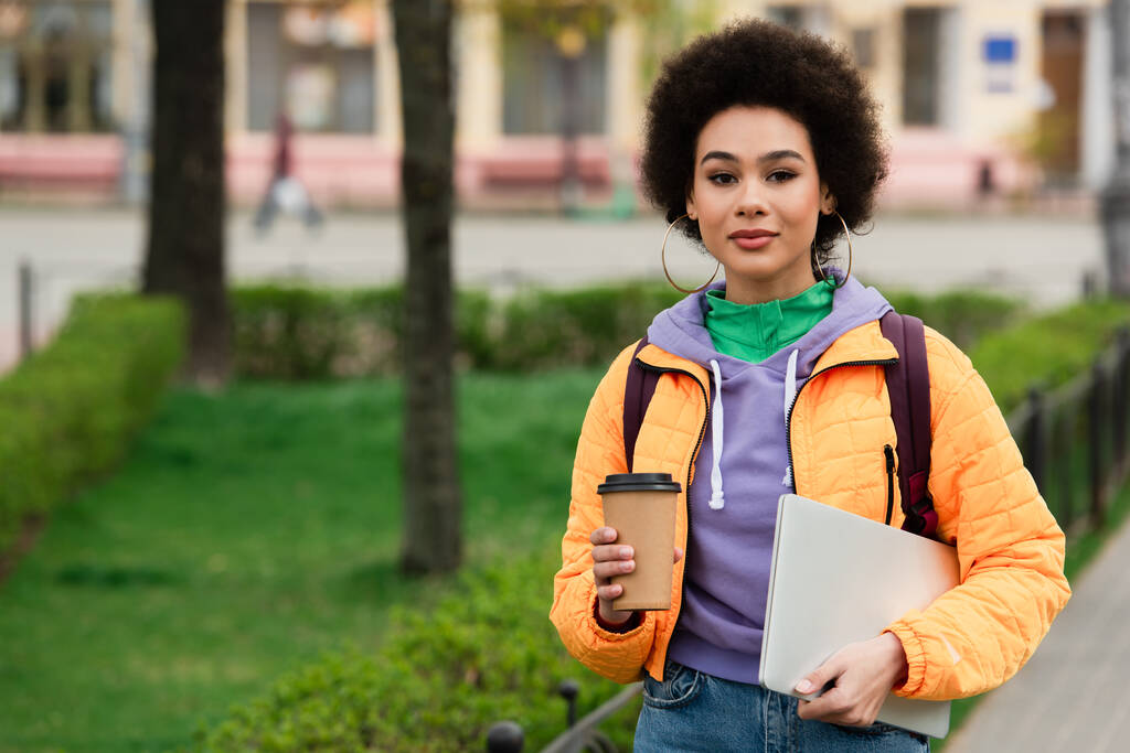Африканская американка смотрит в камеру, держа в руках бумажную чашку и ноутбук на городской улице  - Фото, изображение