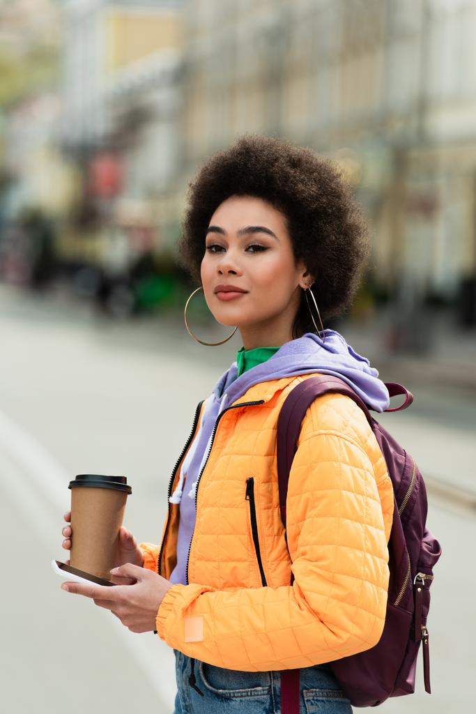 Młoda Afroamerykanka z plecakiem, papierową filiżanką i smartfonem patrząca na kamerę na zewnątrz  - Zdjęcie, obraz