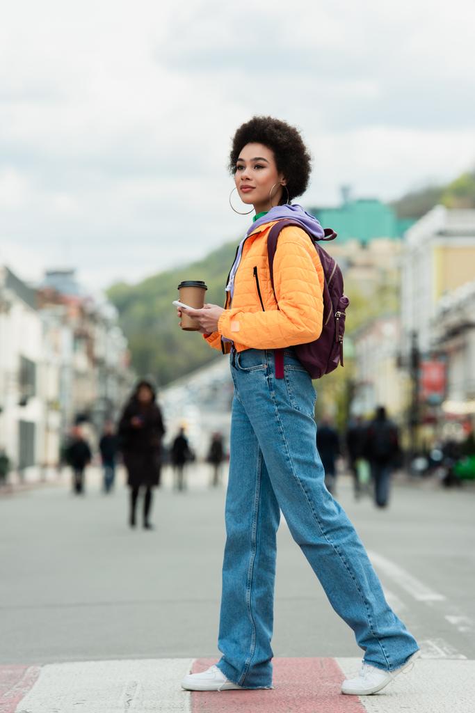 アフリカ系アメリカ人の女性がコーヒーを飲みに街をスマホで歩く  - 写真・画像