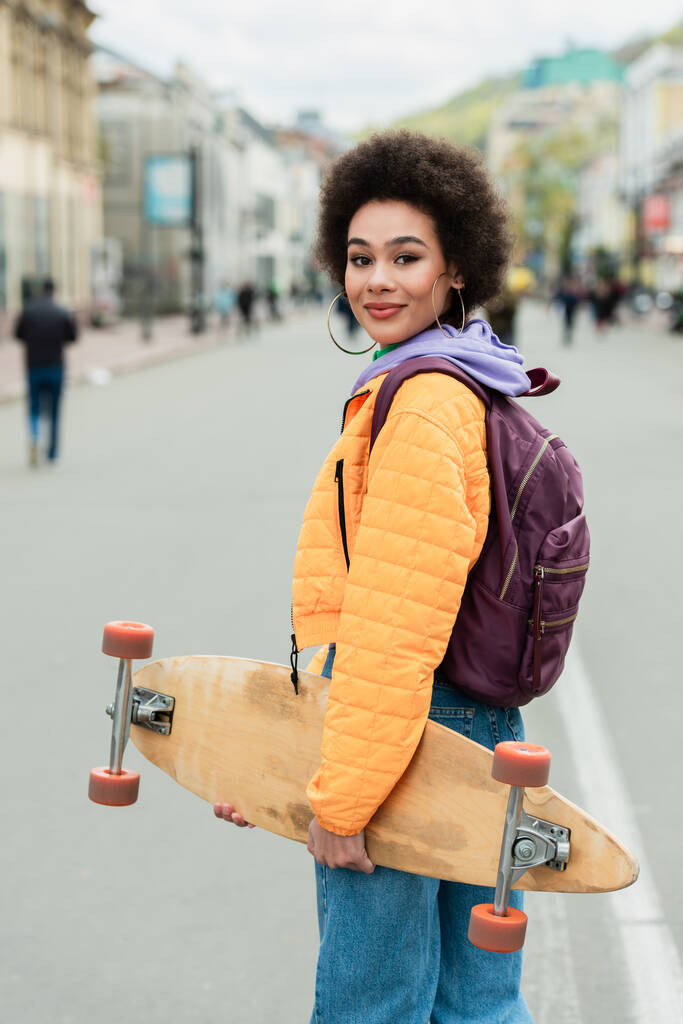 Sorrindo mulher afro-americana com mochila e longboard de pé na rua urbana  - Foto, Imagem