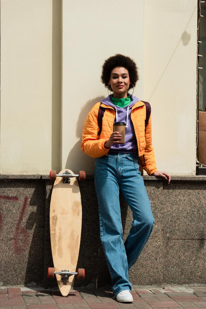 Kağıt bardaklı gülümseyen Afro-Amerikan kadın bina ve uzun tahtanın yanındaki kameraya bakıyor.  - Fotoğraf, Görsel