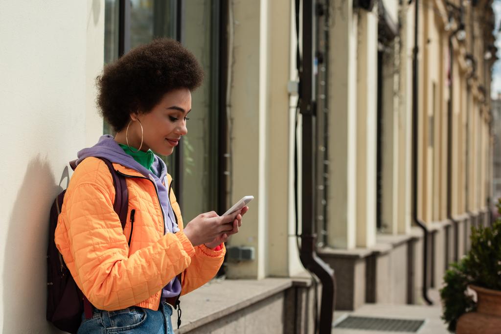 Sonriente mujer afroamericana con mochila usando teléfono inteligente cerca del edificio  - Foto, imagen