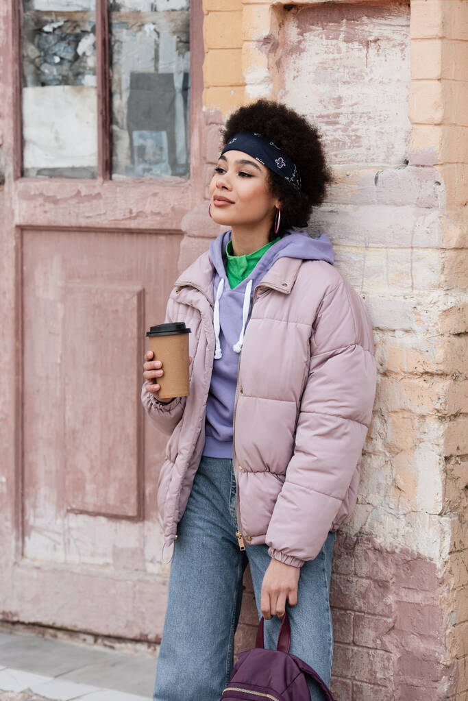 Ładna Afroamerykanka z plecakiem trzymająca kawę w pobliżu starego budynku  - Zdjęcie, obraz