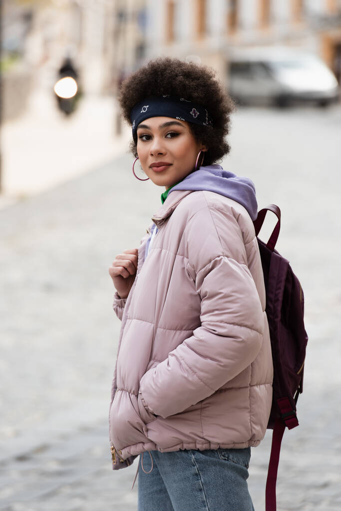 Mujer afroamericana con chaqueta sosteniendo mochila en calle urbana  - Foto, imagen