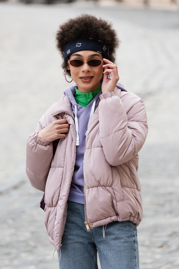 Африканская американка в солнечных очках держит рюкзак и улыбается на городской улице  - Фото, изображение