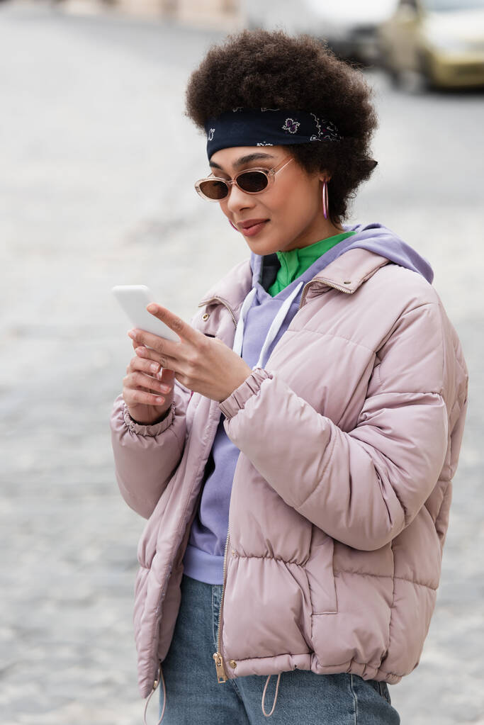 Afroamerikanerin mit Kopftuch und Sonnenbrille nutzt Smartphone im Freien  - Foto, Bild