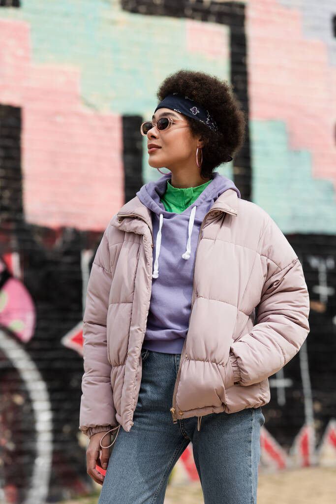 Mujer afroamericana en gafas de sol cogidas de la mano en el bolsillo de la chaqueta en la calle urbana  - Foto, imagen