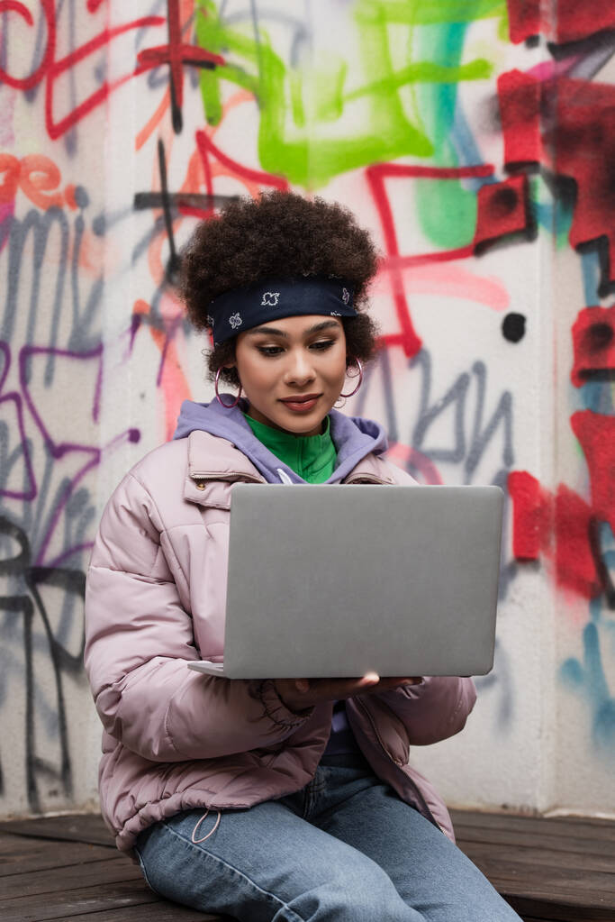 Joven mujer afroamericana usando laptop cerca de graffiti al aire libre  - Foto, imagen
