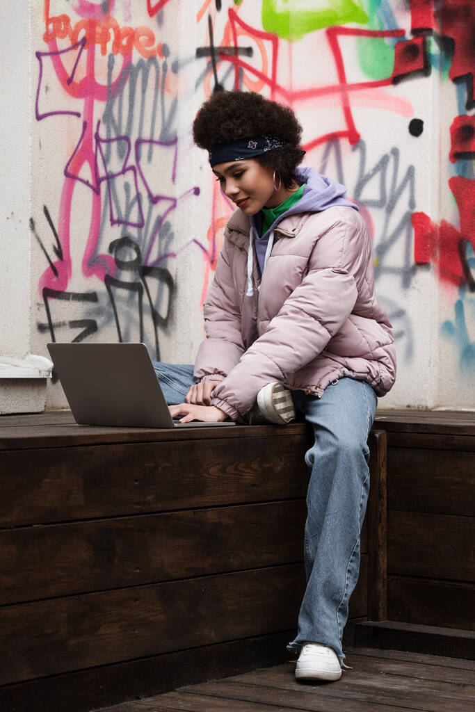 Africano americano teleworker usando laptop no banco perto de graffiti ao ar livre  - Foto, Imagem