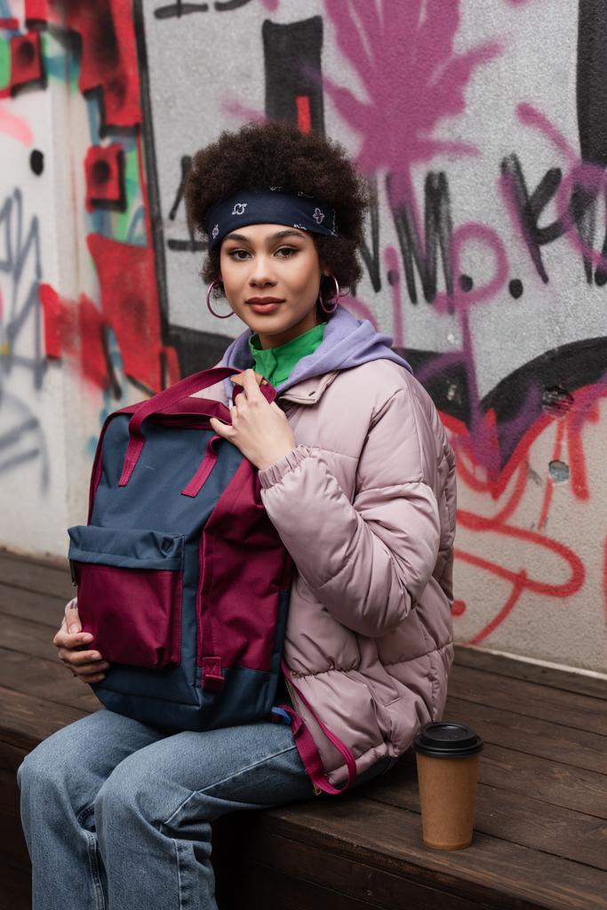 Afričanky americká žena drží batoh u papírového kelímku na lavičce a graffiti - Fotografie, Obrázek