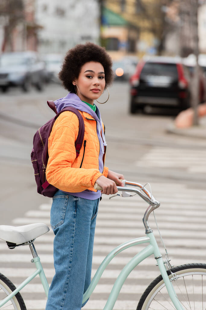 Afroamerikanerin mit Rucksack steht neben Fahrrad auf Stadtstraße  - Foto, Bild