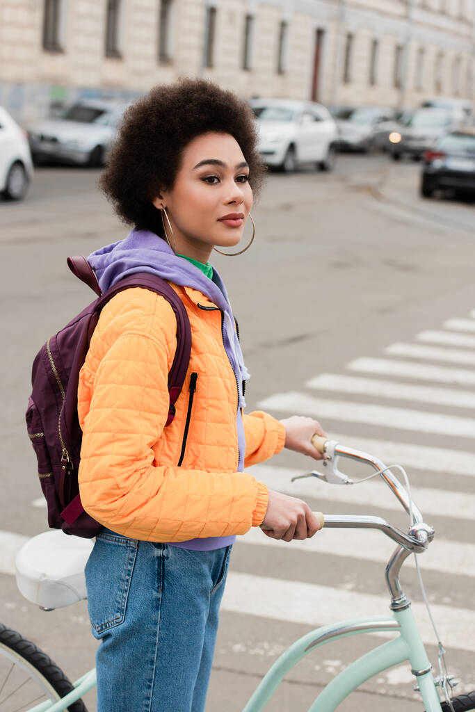 Mujer afroamericana con bicicleta mirando hacia otro lado cerca de paso peatonal borroso al aire libre  - Foto, Imagen