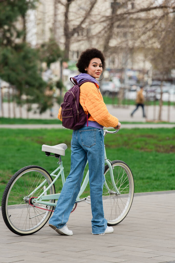African american woman with backpack looking at camera near bike on urban street  - Photo, Image