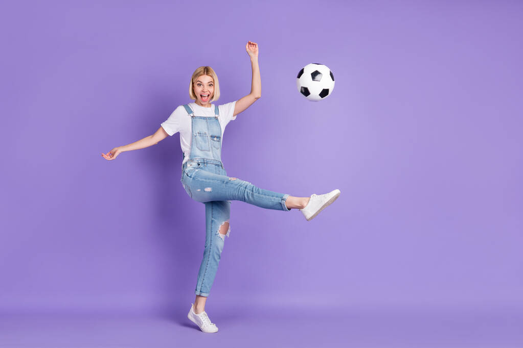 Полноразмерная фотография девушки, счастливой позитивной улыбкой, играющей в футбол с мячом, изолированной над фиолетовым цветом фона - Фото, изображение