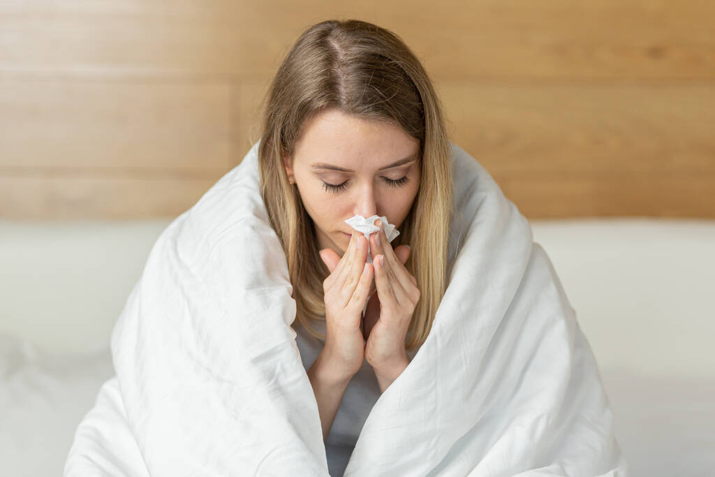 Genç hasta bir kadın yatak odasında, battaniyeye sarılı bir şekilde yatarken burnunu peçeteye sümkürdü. Soğuk kadın ya da kız ya da evde grip. Yakın plan portre Çekici genç sarışın - Fotoğraf, Görsel