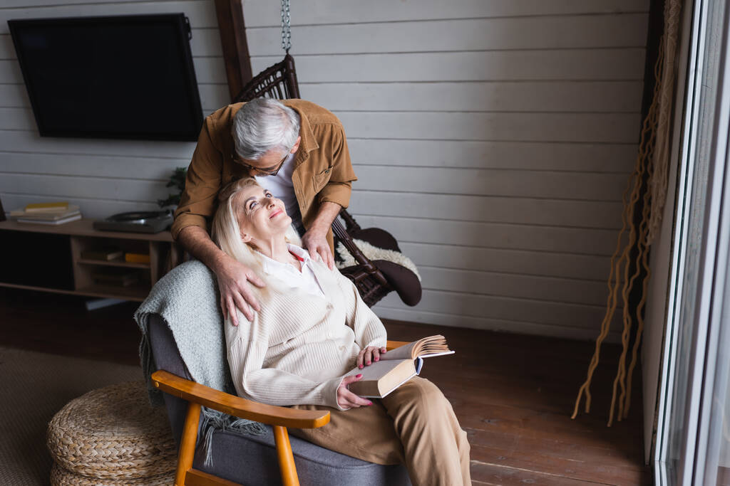 Idős férfi ölelés mosolygós feleség könyvvel karosszékben otthon  - Fotó, kép