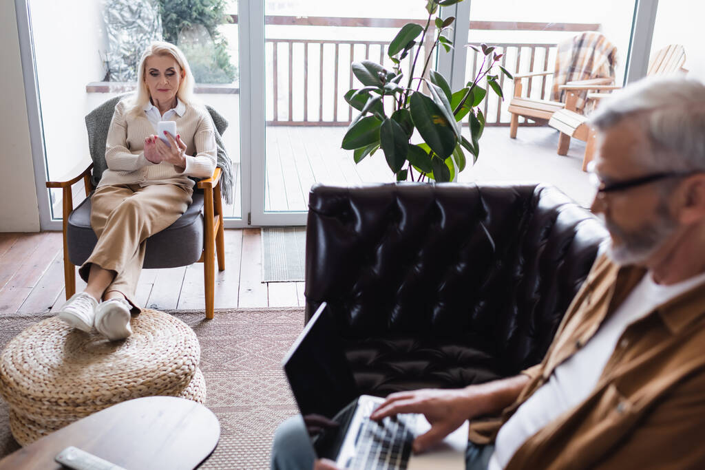 Старша жінка використовує смартфон біля розмитого чоловіка з ноутбуком на дивані
  - Фото, зображення