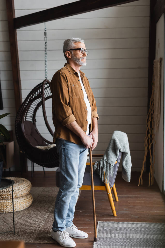 Starší muž v brýlích drží berlu, zatímco stojí doma  - Fotografie, Obrázek