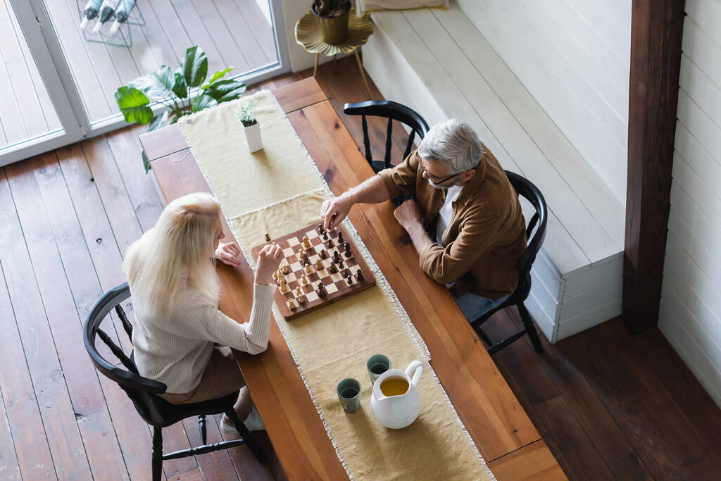 Aus der Vogelperspektive: Senior-Paar spielt Schach in der Nähe von Krug und Gläsern auf Tisch  - Foto, Bild