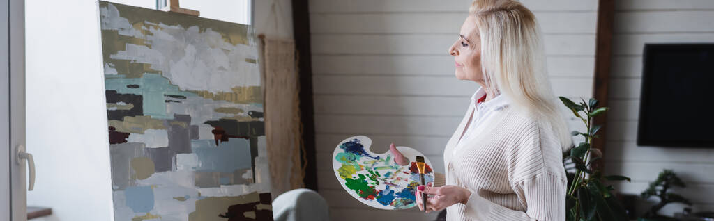 Seitenansicht der Seniorin mit Palette und Pinsel in der Nähe der Malerei, Banner  - Foto, Bild