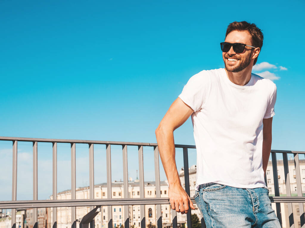 Portret przystojnego uśmiechniętego hipstera lamberseksualnego modelki.Tylowy mężczyzna ubrany w białą koszulkę. Moda mężczyzna pozowanie za niebieskim niebem na tle ulicy w okularach przeciwsłonecznych - Zdjęcie, obraz