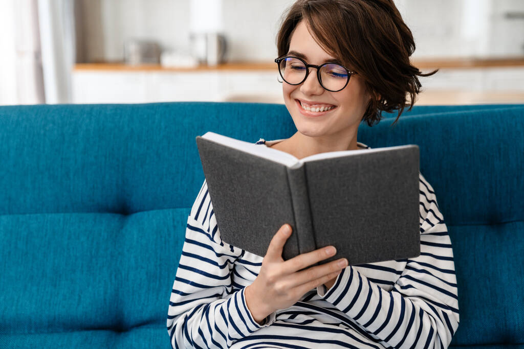Щаслива красива жінка в окулярах читає книгу, сидячи на дивані вдома
 - Фото, зображення