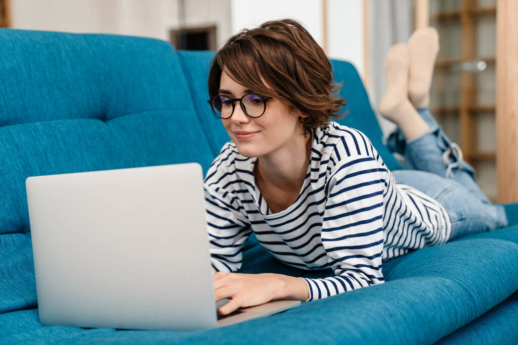 Szczęśliwa piękna kobieta w okularach za pomocą laptopa podczas leżenia na kanapie w domu - Zdjęcie, obraz