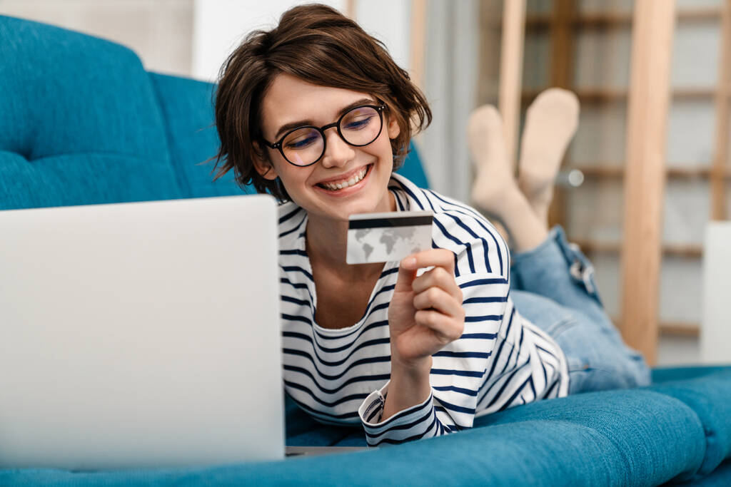 Joyeux belle femme dans les lunettes en utilisant la carte de crédit et ordinateur portable sur le canapé à la maison - Photo, image