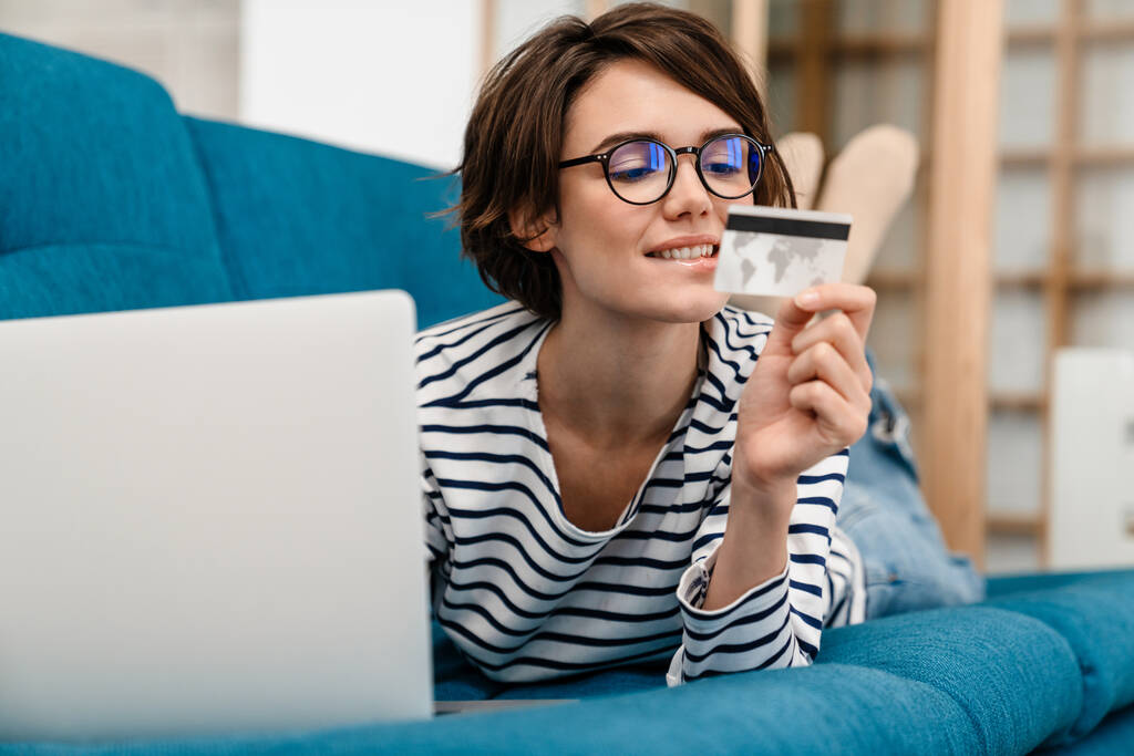 Happy beautiful woman in eyeglasses using credit card and laptop on couch at home - Photo, Image