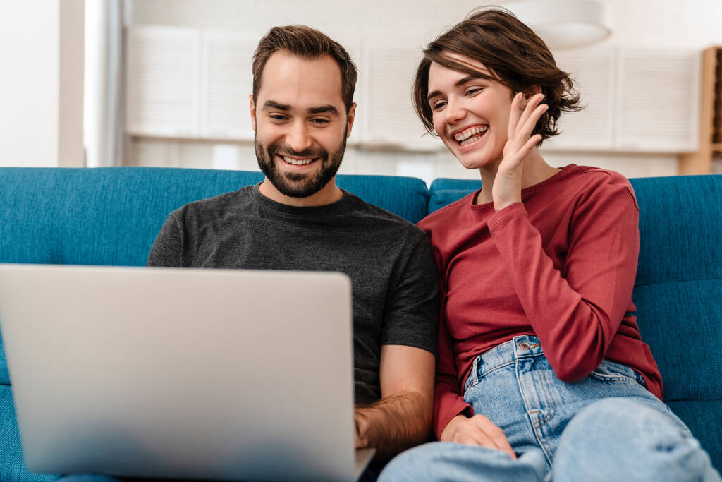 Feliz pareja joven saludando de la mano y el uso de la computadora portátil mientras está sentado en el sofá en casa - Foto, Imagen
