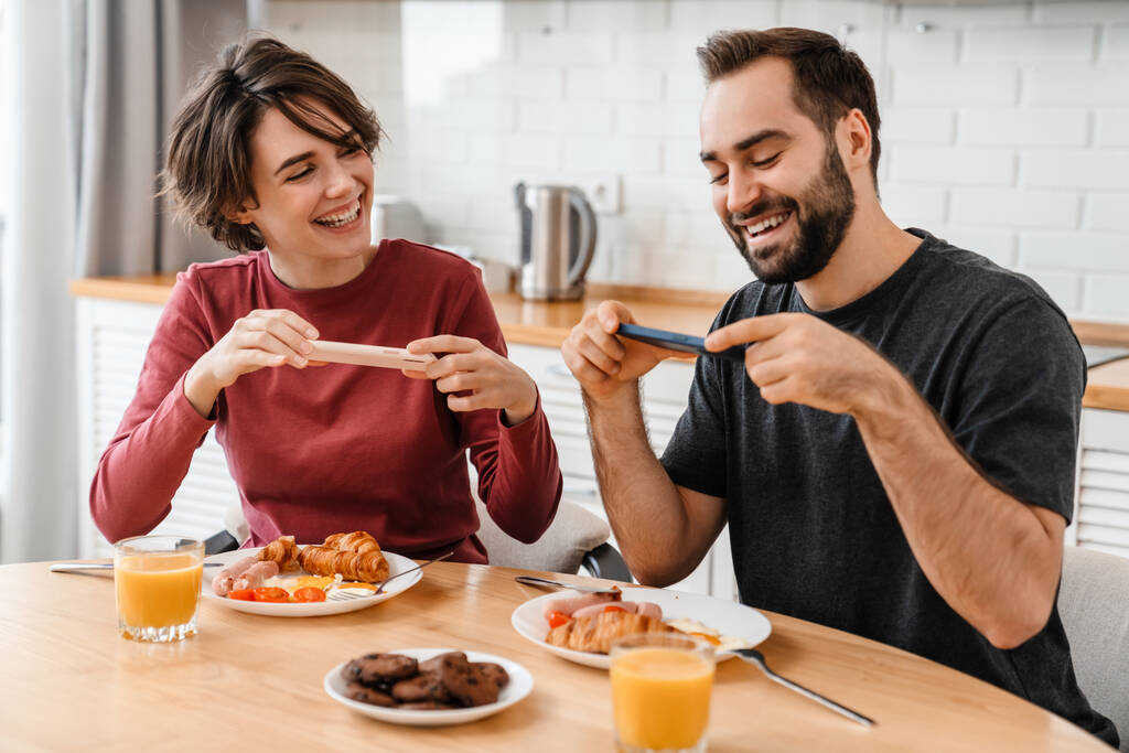 Portrait de jeune couple caucasien prenant une photo sur téléphone portable tout en prenant le petit déjeuner dans une cuisine confortable à la maison - Photo, image