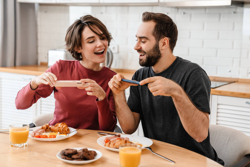 Ritratto di giovane coppia caucasica che scatta foto sul cellulare mentre fa colazione in una cucina accogliente a casa - Foto, immagini