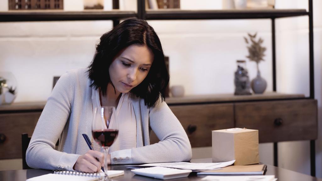 σοβαρή γυναίκα κάθεται στο τραπέζι με ένα ποτήρι κόκκινο κρασί και γράφοντας στο σημειωματάριο στο σπίτι - Φωτογραφία, εικόνα