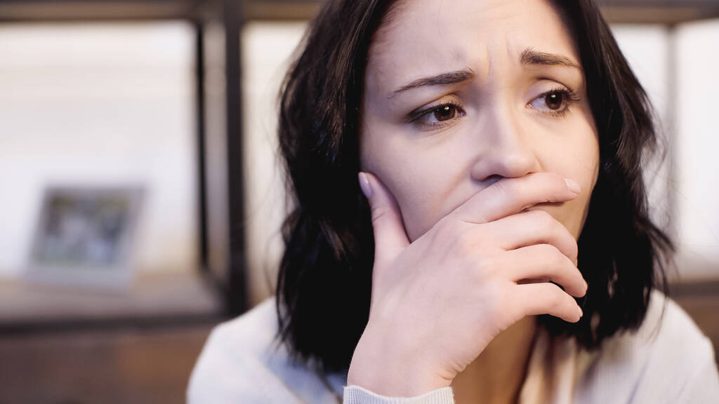 портрет депрессивной брюнетки в бежевом свитере, покрывающем рот рукой дома - Фото, изображение