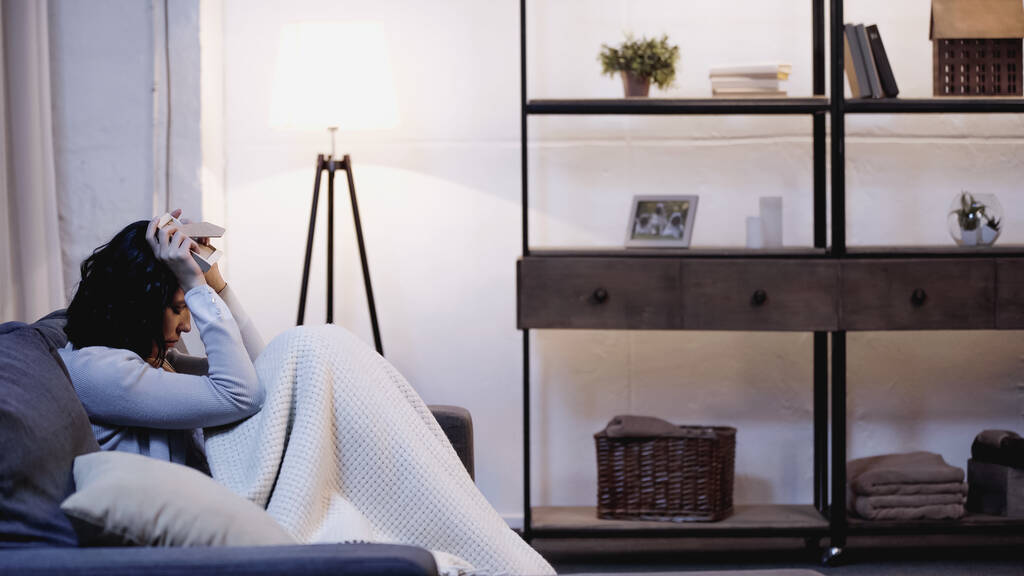 Depressive Frau in Decke gehüllt sitzt auf Sofa und hält Fotorahmen in Kopfnähe zu Hause - Foto, Bild