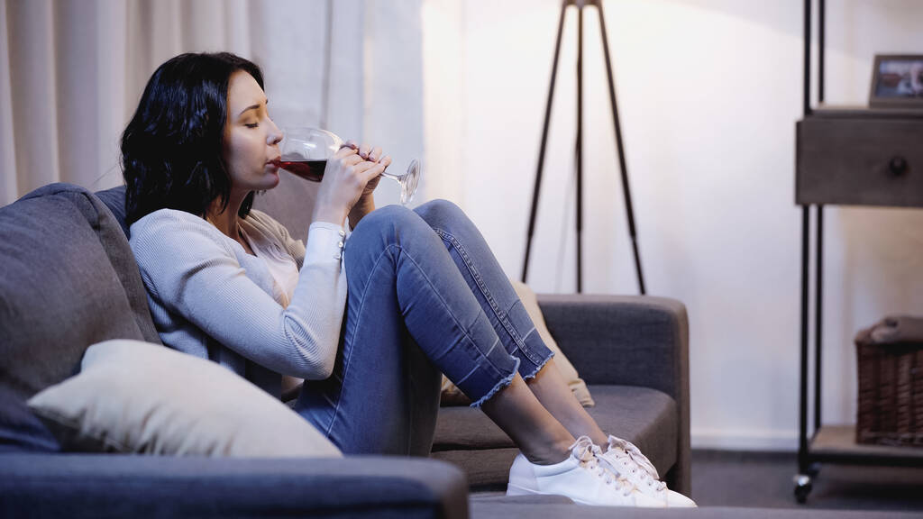 mujer deprimida y solitaria en ropa casual sentada en el sofá y bebiendo vino tinto de vidrio en casa - Foto, imagen
